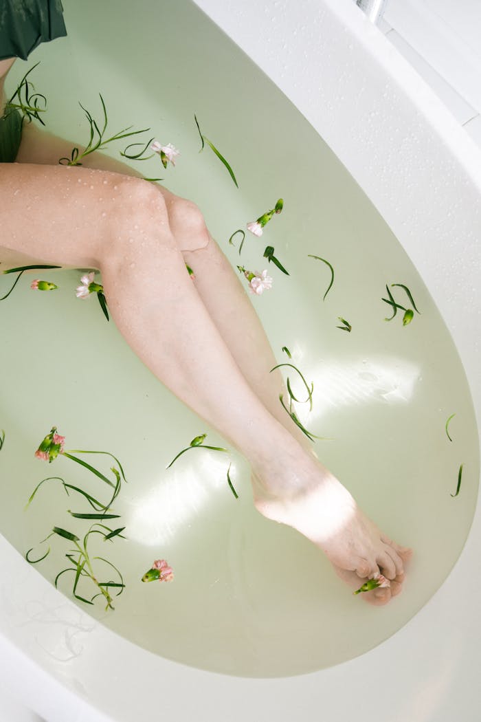 A serene top view of legs in a bathtub with floating flowers and greenery.